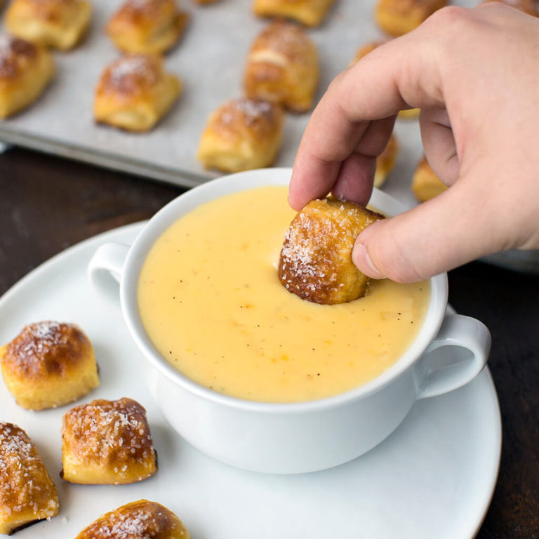 Hand dipping a freshly baked pretzel bite into a creamy cheese dip on a white plate, perfect for appetizers and snacks.