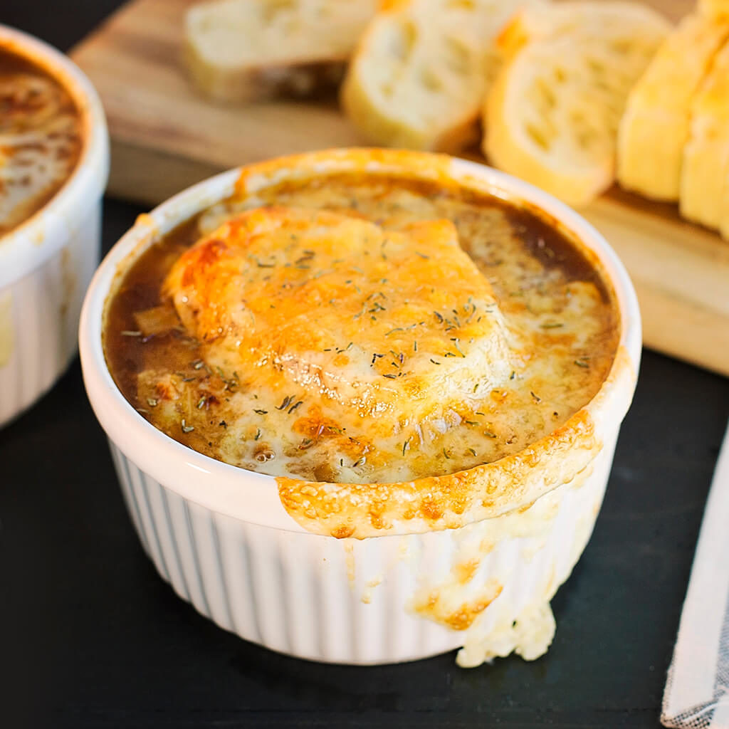 French onion soup in white bowl topped with melted cheese and thyme, served with bread.