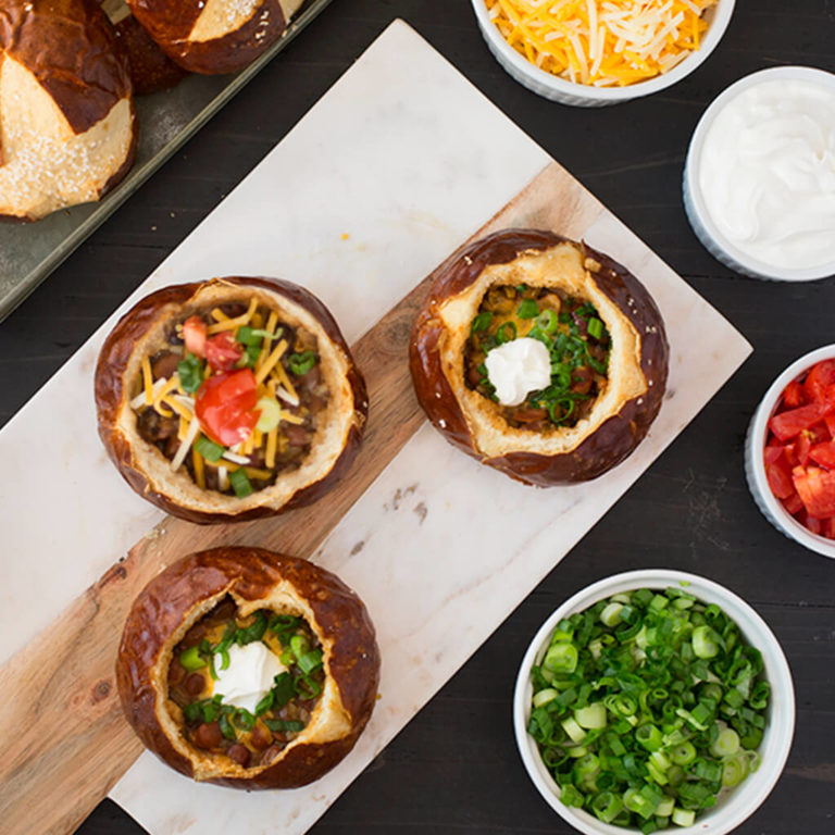 Homemade bread bowls filled with chili, topped with cheese and sour cream, surrounded by fresh toppings.