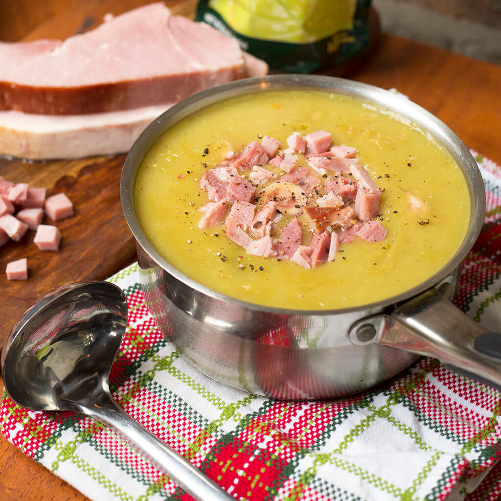 Creamy pea soup garnished with diced ham in a silver pot, served with bread on a rustic table.